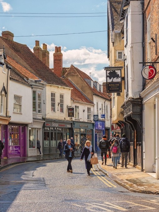 Shopping street just after a rain shower. Historic buildings line the street and shoppers walk between them. A sky with cloud is overhead.