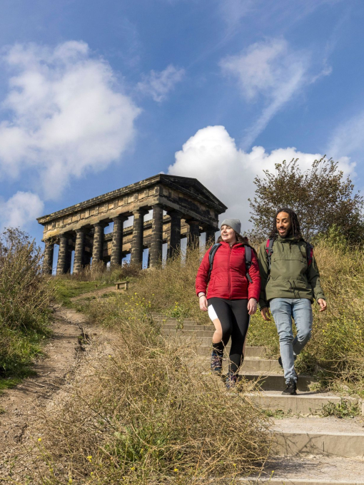 Two people wearing outdoor clothing, hiking past a stone monument, surrounded by greenery and blue skies.