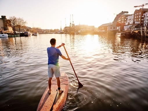 Rear view of a man standing on a paddleboard in a harbour at dawn.