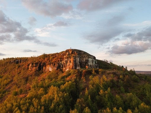 A lone person stands on a cliff top with forest below
