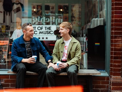 Two men sit with take away drinks in front of a shop