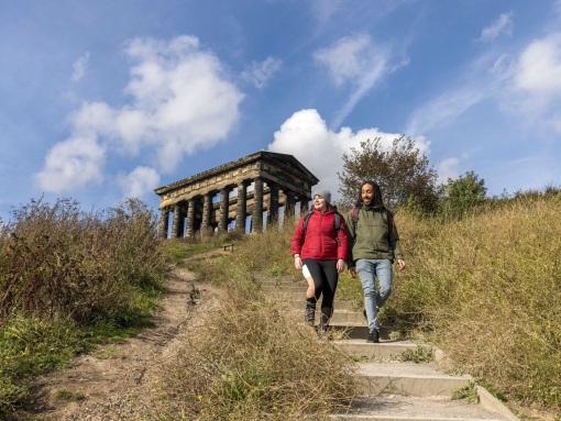 Two people wearing outdoor clothing, hiking past a stone monument, surrounded by greenery and blue skies.
