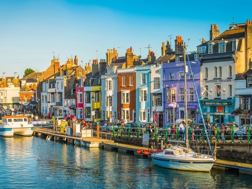 People walking up and down the water front of a busy seaside town lined with colourful terraced houses and boats anchored in the harbour