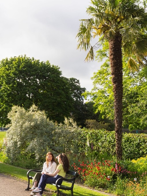 Two women sit on a bench in public gardens