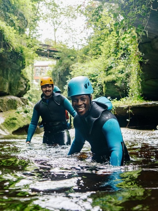 Two men wearing wetsuits and helmets wade waist deep in a gorge