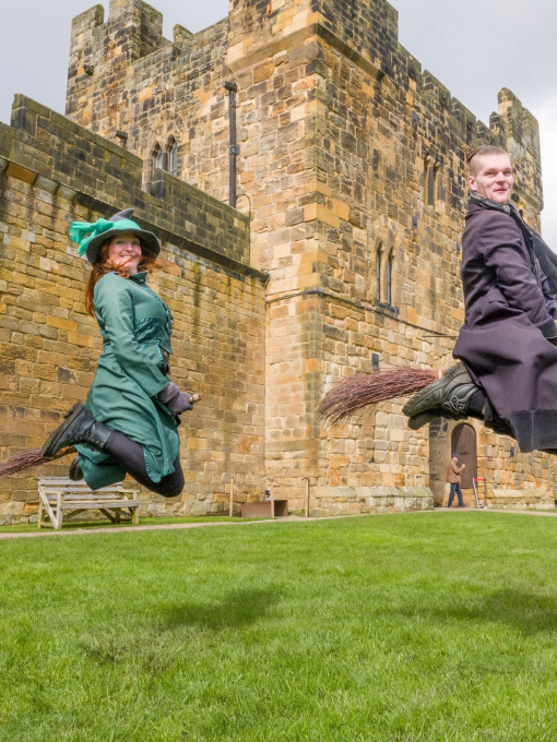Two wizarding professors demonstrating flying on a broom outside a large castle.