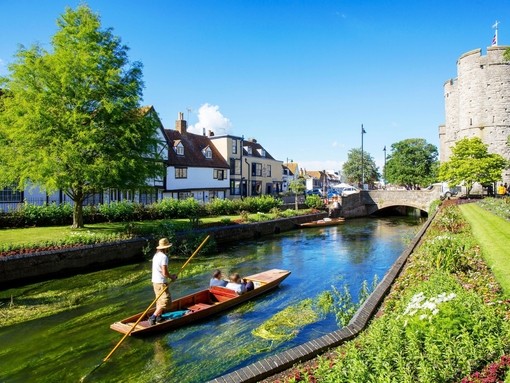 A man and a couple in a punt on a small river surrounded by gardens and houses