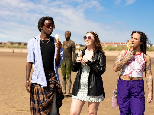 A group of friends walk on the beach with ice creams, a statue in the background