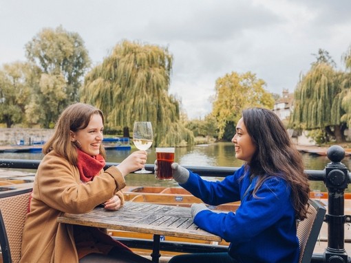 Two women toasting with drinks at a pub beside the river