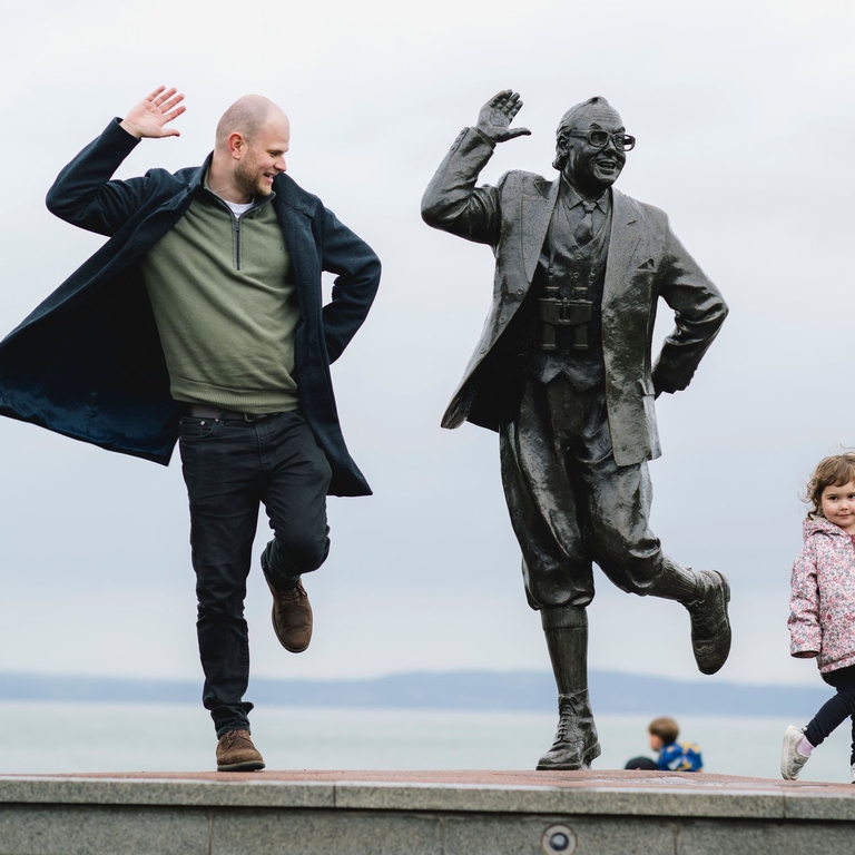 View of man, female child and statue of a man in front of the sea